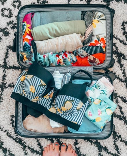 Suitcase packed with beach supplies, a lemon-printed bikini, and heart-shaped sunglasses on a black and white patterned carpet.