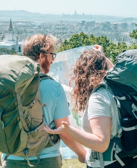 Couple wearing heavy backpacks looking confused at a map in front of a city skyline