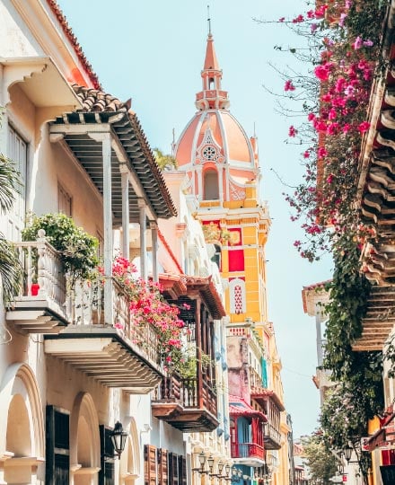 Flower-covered balconies in the foreground lining a street, with the yellow and pink tower of Cartagena, Colombia's cathedral rising above.