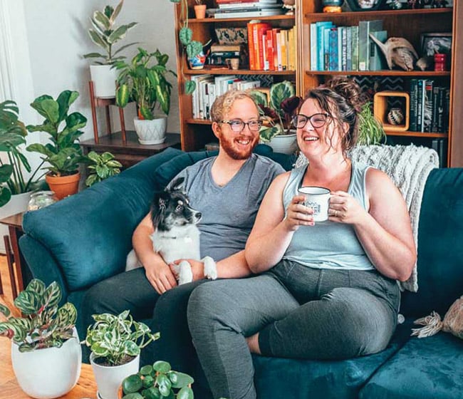 Couple on a blue couch surrounded by plants, holding a fluffy black and white dog and laughing